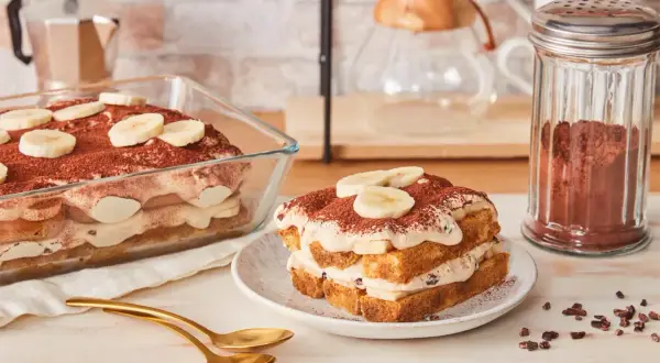 Ein köstliches Bananen Tiramisu, geschichtet mit Creme und Bananenscheiben, bestäubt mit Kakao. Daneben ein Glas Kakaopulver.