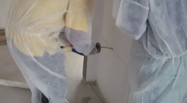 Dos operarios con trajes protectores aplican espuma intumescente en una pared, sellando una junta para protección contra incendios.