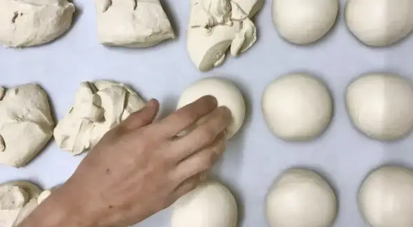 Des boules de pâte à pizza sont disposées sur une plaque. Une main façonne délicatement l'une d'elles.