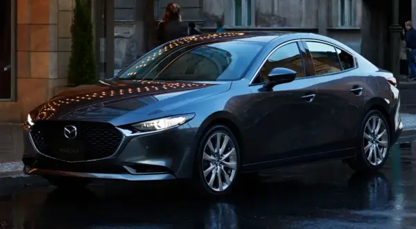 Elegancka, ciemnoszara Mazda 3 sedan stoi na mokrej ulicy, odbijając światła miasta.