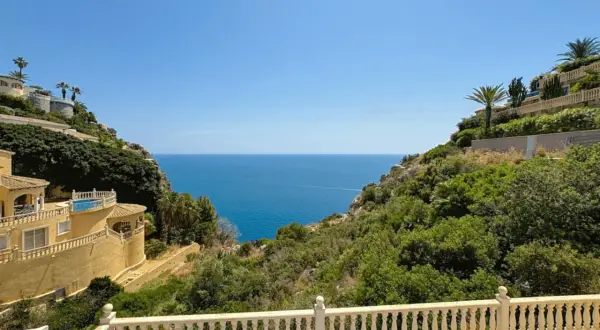 Vista desde una terraza con barandilla blanca hacia el mar azul, con villas en laderas verdes. La puerta del mar San José se abre a un paisaje idílico.