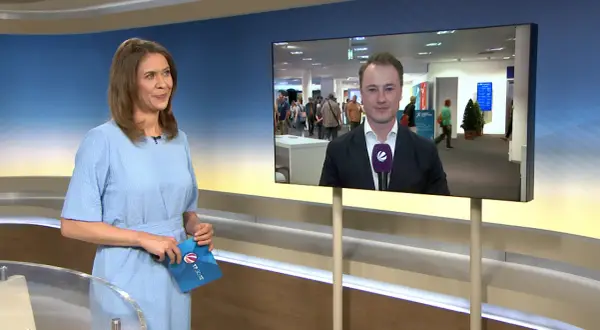 Moderatorin hält Umschlag mit Logo von tv-n hessen. Auf dem Bildschirm ein Reporter im Anzug.