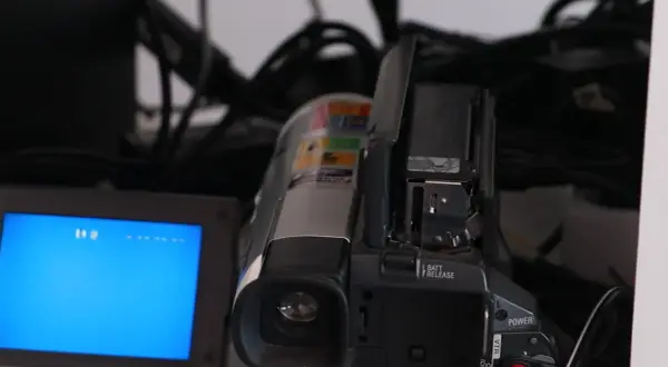 En videokamera med öppen lucka och en blå skärm. Kablar syns i bakgrunden, en påminnelse om analoga bilder och äldre teknik.