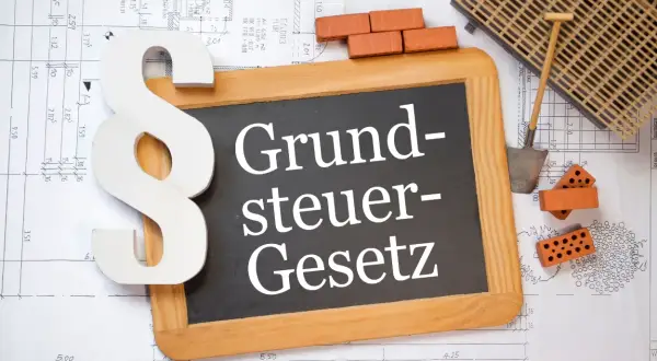 Ein Paragrafensymbol und eine Tafel mit "Grundsteuer-Gesetz" liegen auf Bauplänen. Ziegel und Werkzeug deuten auf den Grunderwerbsteuer in Niedersachsen hin.