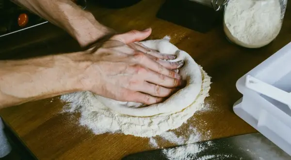 Dłonie delikatnie rozciągają ciasto na pizze, tworząc idealny okrąg na drewnianym blacie. Mąka rozsypana wokół sugeruje, jak rozciągnąć ciasto na pizze.