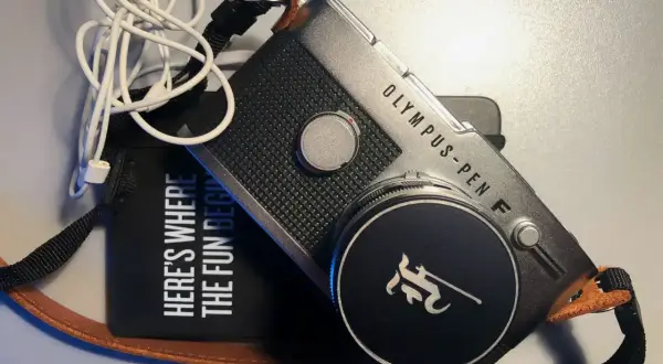 The Olympus Pen FV camera, with its iconic design, sits beside a black pouch that reads "HERE'S WHERE THE FUN BEGINS." White earbuds are tangled nearby.