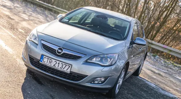 Srebrny Opel Astra J z przodu, zaparkowany na drodze z drzewami w tle.