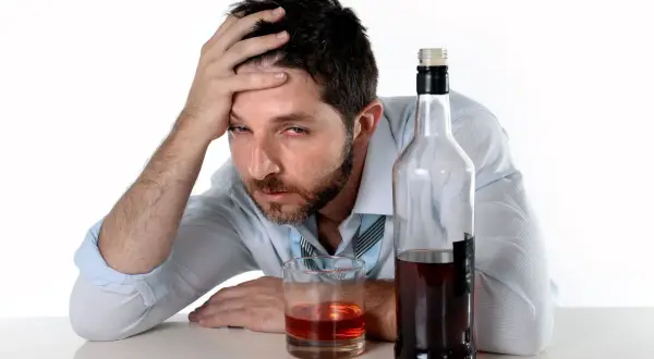Mężczyzna z ręką na czole obok butelki i szklanki z alkohoholem.