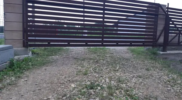 Brązowa, metalowa brama przesuwna z poziomymi szczeblinami. Widoczna jest szczelina pod bramą, przez którą widać trawę i żwir.