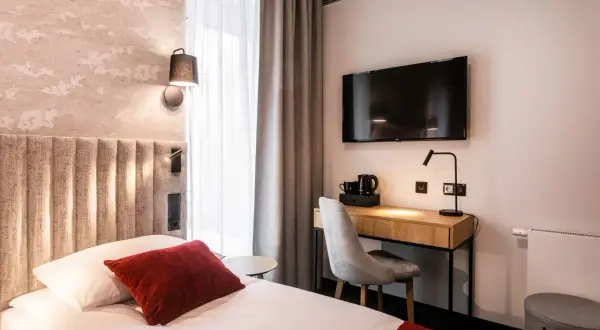 Przytulny pokój w hotelu Vesper House: łóżko z czerwonymi akcentami, biurko z czajnikiem i telewizorem, szare zasłony.