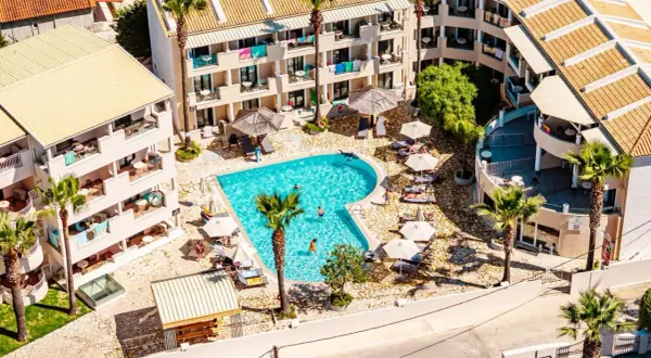 Widok z lotu ptaka na basen i budynki hotelowe. Wiele osób odpoczywa przy basenie. Warto sprawdzić caretta island hotel opinie przed rezerwacją.