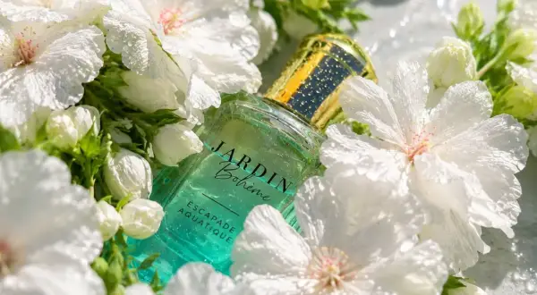 Szklana butelka perfum Jardin Bohème Escapade Aquatique, otoczona białymi kwiatami. Ten zapach to jak szczęśliwy los na lato.