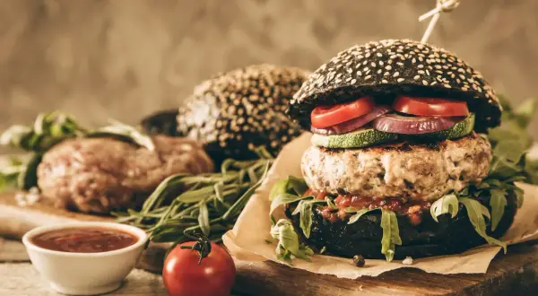 Gotowy hamburger z czarną bułką, kotletem wołowym i warzywami, obok surowy kotlet i sos.