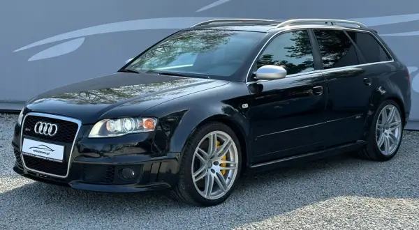 Czarny Audi RS4 B7 Avant z żółtymi zaciskami hamulcowymi, zaparkowany na żwirze przed szarą ścianą z grafiką.