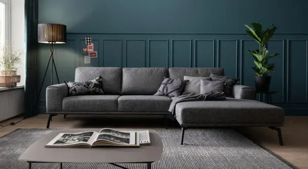 Ein modernes Wohnzimmer mit einem grauen Sofa, das mit Kissen und einer Decke dekoriert ist. Ein Stehlampen-Design und eine Zimmerpflanze runden das Bild ab.