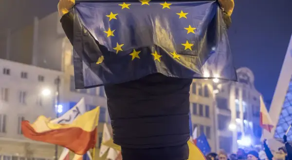 Osoba trzyma flagę UE, symbol jedności, która powstała w 1993 roku. W tle widać inne flagi i tłum ludzi.