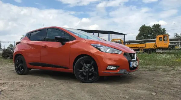 Pomarańczowy Nissan Micra na tle torów kolejowych. Sprawdź nissan micra wymiary, by poznać jego kompaktowe gabaryty.