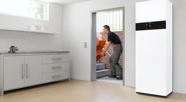 Instalator wnosi nowy, pomarańczowy element, który może być częścią odpowiedzi na pytanie: jaki kocioł gazowy do domu? Obok stoi nowoczesny, biały kocioł.