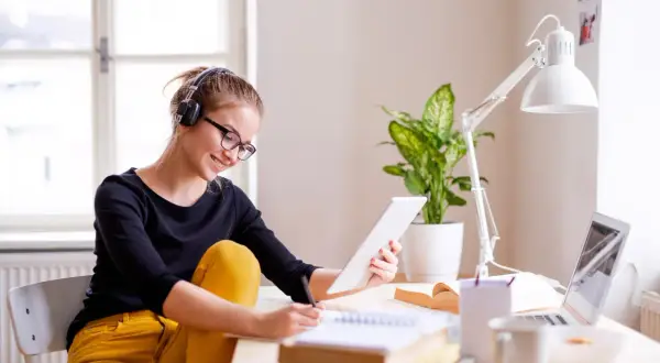 Junge Frau mit Kopfhörern und Brille lernt am Tablet für ihr **vollzeit studium**. Sie schreibt Notizen in ein Heft.
