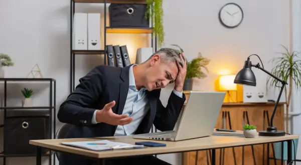 Zestresowany mężczyzna w garniturze trzyma się za głowę, patrząc na laptopa. Może właśnie stracił wadium.