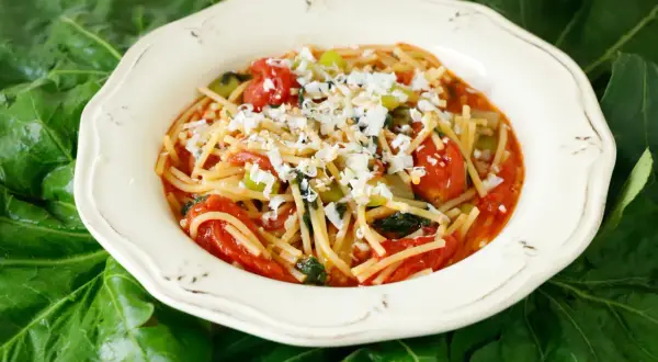 Un piatto di pasta con i tenerumi asciutta, condita con pomodoro fresco e formaggio grattugiato, adagiato su foglie verdi.