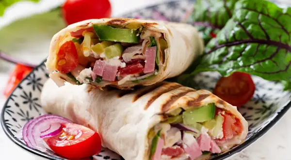 Pyszna tortilla z szynką, awokado, pomidorami i serem feta, podana z sałatką i czerwoną cebulą.