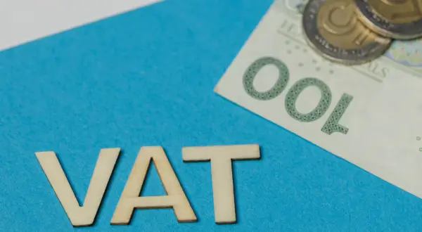 Drewniane litery VAT na niebieskim tle, obok banknot 100 zł i monety. Rozważanie vat należny a naliczony.