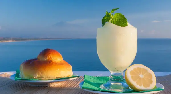 Colazione siciliana con brioche e granita al limone, con il mare e l'Etna sullo sfondo.