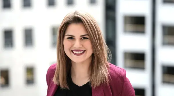 Lächelnde junge Frau mit braunen Haaren, die einen pinken Blazer trägt. Sie studiert IU Master Wirtschaftsinformatik und blickt optimistisch in die Zukunft.