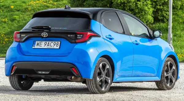 Niebieski Toyota Yaris IV z czarnym dachem, zaparkowany na żwirowej drodze, z zielonym tłem.