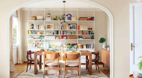 Comedor con decoración estilo nórdico, mesa de madera, sillas de diseño y estanterías blancas repletas de libros y objetos decorativos.