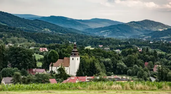 Widok na wieś z kościołem i domami, otoczoną przez zielone wzgórza i lasy. W oddali widać łagodne **górki wielkie**.