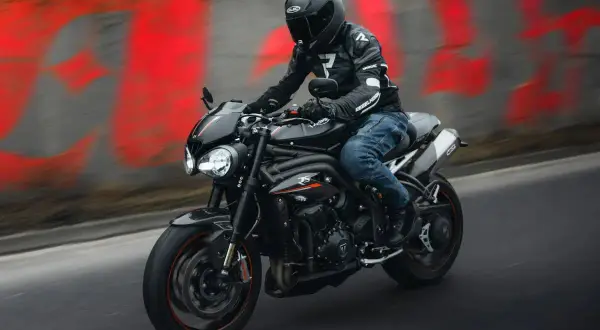 Motocyklista w kasku i kurtce jedzie czarnym motocyklem na tle czerwonych napisów.