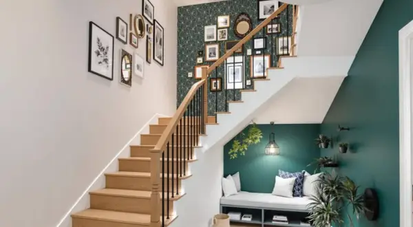 Un habillage mural de cage d'escalier avec une galerie de cadres et un papier peint géométrique vert. Un banc confortable est aménagé sous l'escalier.