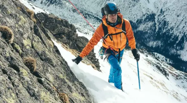 Alpinista w pomarańczowej kurtce wspina się po skalistym, ośnieżonym zboczu, używając czekana alpinisty inaczej jako wsparcia.