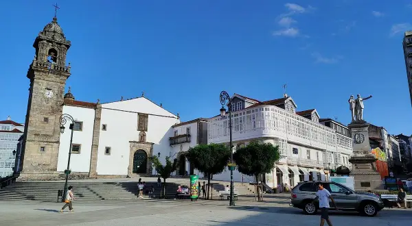 La plaza Betanzos, con su iglesia de torre campanario, edificios blancos y un monumento, bajo un cielo azul.