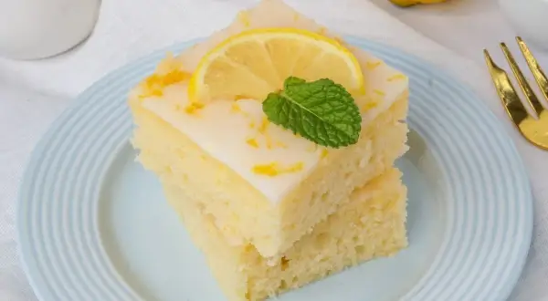 Zwei Stücke zitronenkuchen mit joghurt, garniert mit Zitronenscheibe und Minze, auf einem hellblauen Teller.