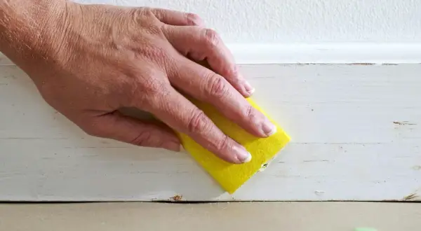 Hand med gul svamp slipar en vitmålad list. Förberedelser för att måla tunt.