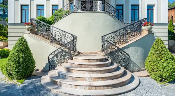 Imponente villa con scale esterne coperte da ringhiere in ferro battuto e ornate da vasi con fiori rossi.