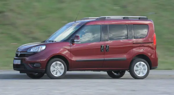 Czerwone kombi auto w ruchu, z widocznymi drzwiami przesuwnymi i relingami dachowymi, na tle rozmytego zielonego krajobrazu.