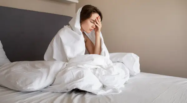 Kobieta w łóżku, przykryta pościelą, z ręką na czole. Wygląda na zmęczoną i zmartwioną, być może przez koszmary w ciąży.