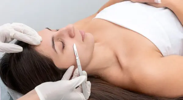 Zabieg dermaplaningu: kosmetyczka w rękawiczkach delikatnie usuwa martwy naskórek i włoski z twarzy klientki za pomocą skalpela.