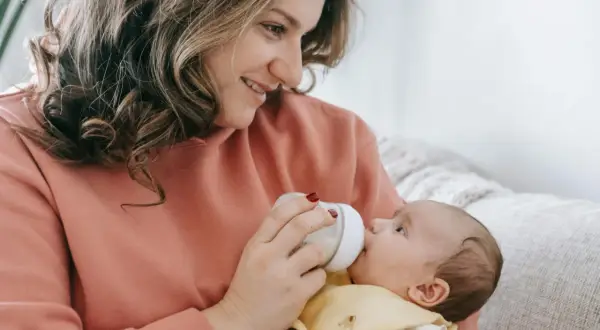 Uśmiechnięta mama karmi butelką dziecko. Warto wiedzieć, ile odciągnięte mleko może stać w lodówce, by zapewnić mu bezpieczeństwo.