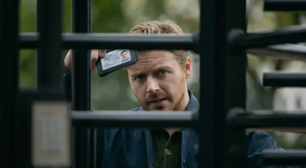 Ein Mann mit blondem Haar und Bart hält einen Ausweis hoch, während er durch ein Gitter blickt. Spannung in Slow Horses Staffel 3.