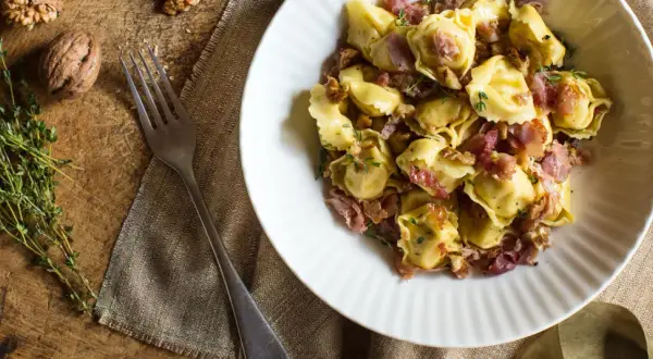 En skål med tortellini prosciutto, valnötter och timjan. En gaffel ligger bredvid på en rustik träyta.