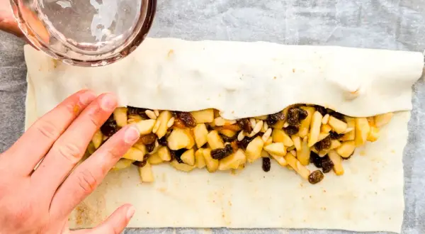 Mani che chiudono uno strudel di mele con pasta sfoglia, ripieno di mele a cubetti, uvetta e pinoli.