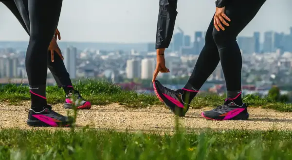Zwei Läuferinnen dehnen sich auf einem Hügel mit Blick auf eine Stadt. Sie tragen schwarze Laufhosen und auffällige pink-schwarze Laufschuhe. Ein Zeichen für den Wiedereinstieg ins joggen nach Geburt.