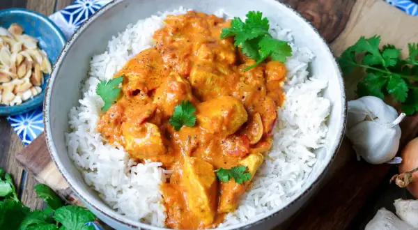 Schüssel mit duftendem Reis und cremigem Chicken Tikka, garniert mit frischem Koriander. Daneben Mandelsplitter, Knoblauch und Ingwer.