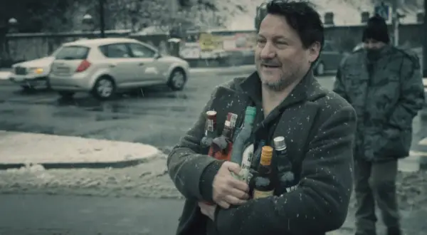 Mężczyzna z uśmiechem niesie kilka butelek alkoholu pod mocnym aniołem obsada. W tle śnieg i samochody.