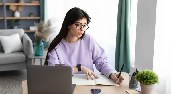 Kobieta w okularach pisze w zeszycie, szukając informacji, jak znaleźć swój numer IBAN. Obok leży laptop i telefon.